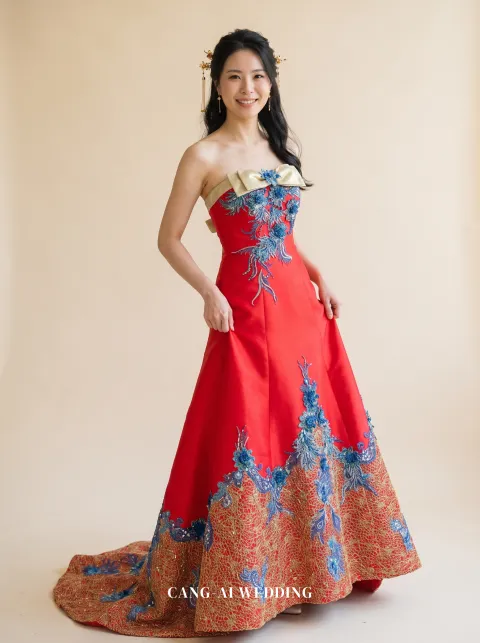 Front full-length of Cuiwei, a strapless red A-line gown with a gold band and blue embroidered hem, lifting the skirt slightly at the hips, reference 6-NR10.