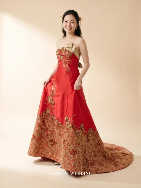 Front full-length of Jincheng, a strapless red satin A-line kua with a gold neckline band and gold-embroidered hem, lifting the skirt slightly at the hips, reference 16-NR05.